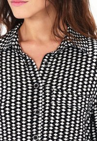 La camisa de patrón blanco y negro presenta un diseño de diamantes, un gran cuello, botones y un único bolsillo en el pecho. La textura del tejido suave es visible.