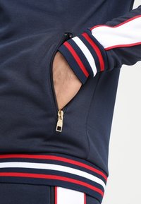 Chaqueta atlética azul marino con un bolsillo con cremallera, puños de canalé a rayas en colores rojo, blanco y azul marino, y una tela suave y texturizada.