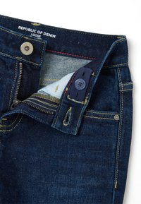 Jeans de mezclilla azul oscuro con un corte holgado. Presentan un cierre de cremallera, costuras amarillas y un cierre de botón con un bolsillo interno. Etiqueta de la marca visible.