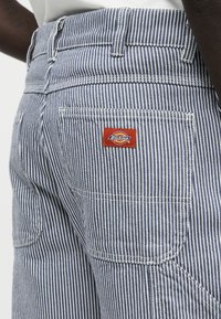 Κοντινή λήψη πίσω τσέπης σε μπλε και λευκό ριγέ παντελόνι με κόκκινο λογότυπο Dickies στην τσέπη.