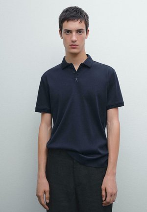 Navy polo shirt with short sleeves and a three-button placket. Smooth cotton fabric with a relaxed fit, paired with dark pants.
