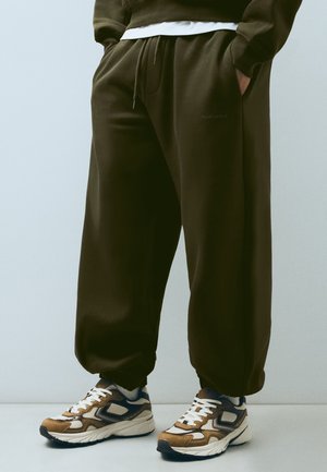 Persona che indossa pantaloni della tuta larghi verde scuro e scarpe da ginnastica bianche, marroni e blu navy, in piedi davanti a uno sfondo semplice.