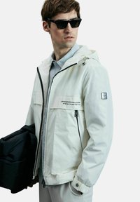 Veste imperméable blanche avec capuche à cordon de serrage, fermeture éclair à l'avant et deux poches zippées. Présente des accents noirs et des détails de logo.