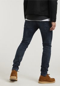 Muž v tmavě modrých slim-fit džínách, hnědých semišových mokasínech, černé bundě a vrstvených černobílých košilích, stojící na neutrálním pozadí.