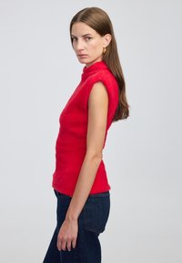 Žena s dlouhými hnědými vlasy, oblečená v bezrukávovém chlupatém červeném topu a tmavě modrých džínách, stojící v profilu na jednobarevném světlém pozadí.