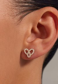 Ohrstecker in Herzform aus Gold mit mehreren kleinen Steinen, die eingearbeitet sind, und verziert mit kunstvoll verschlungenen Schleifen sowie einer polierten Metalloberfläche.