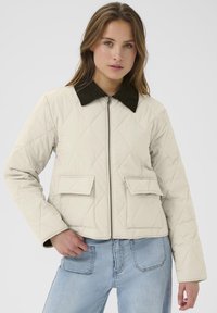 Giacca trapuntata color crema con colletto in velluto a coste nero, chiusura frontale con zip e due tasche a patta. Presenta maniche lunghe e un design corto.