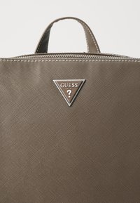 Ruda tekstūruota rankinė su užtrauktuku, su trikampio formos logotipo plokštele, išgraviruota "GUESS" ir klausimo ženklu.