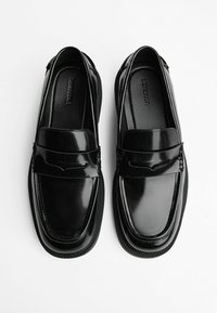 Mocasini din piele patent neagră cu un finisaj neted, accente cusute și o talpă din cauciuc cu modele texturate.