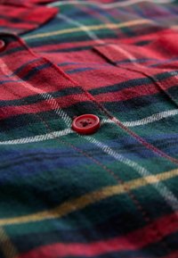 Red and green plaid flannel shirt featuring a prominent red button, soft texture, and a pocket detail in a classic pattern design.