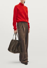Red ribbed high-neck sweater with shoulder buttons, paired with brown wide-leg trousers and a dark brown textured bag. Brown heeled shoes.