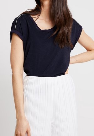 Frau trägt ein kurzärmeliges dunkelblaues V-Ausschnitt-Oberteil, das in hochtaillierte weiße Plisseehosen gesteckt ist, und posiert mit einer Hand in der Hüfte.