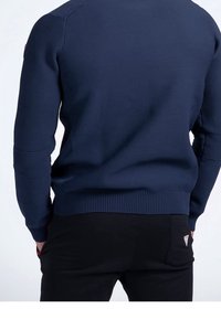 Maglione lavorato a maglia di colore navy con tessuto testurizzato, polsini e orlo a coste, e toppe sulle maniche di colore contrastante. Pantaloni neri con un piccolo logo sulla tasca.
