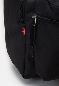 Black backpack made of durable fabric, featuring a zipper pocket and a small red logo tag. The design includes reinforced stitching and textured surface.