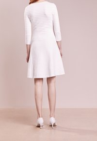 Robe blanche avec un motif texturé, manches trois-quarts, jupe évasée et finition lisse. Associée à des chaussures à talons blancs.