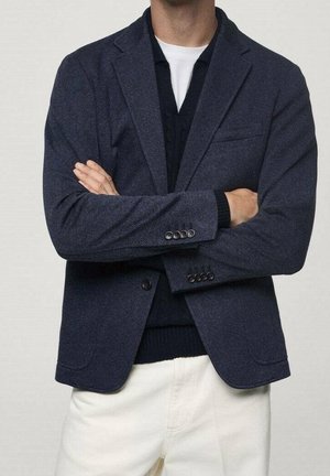 Mann trägt einen dunkelblauen Blazer, dunklen Pullover, weißes T-Shirt und helle Hose mit verschränkten Armen.