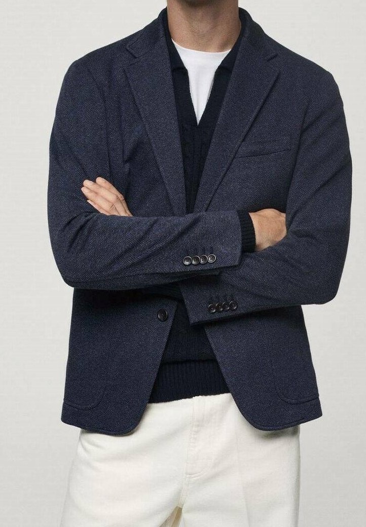 Homme portant un blazer bleu foncé, un pull foncé, un t-shirt blanc et un pantalon clair, les bras croisés.