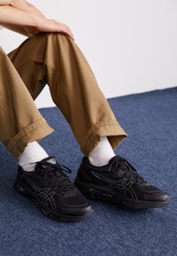 Baskets de course noires avec dessus en mesh, accents réfléchissants et semelle rembourrée, associées à un pantalon beige et des chaussettes blanches montantes sur un tapis bleu.