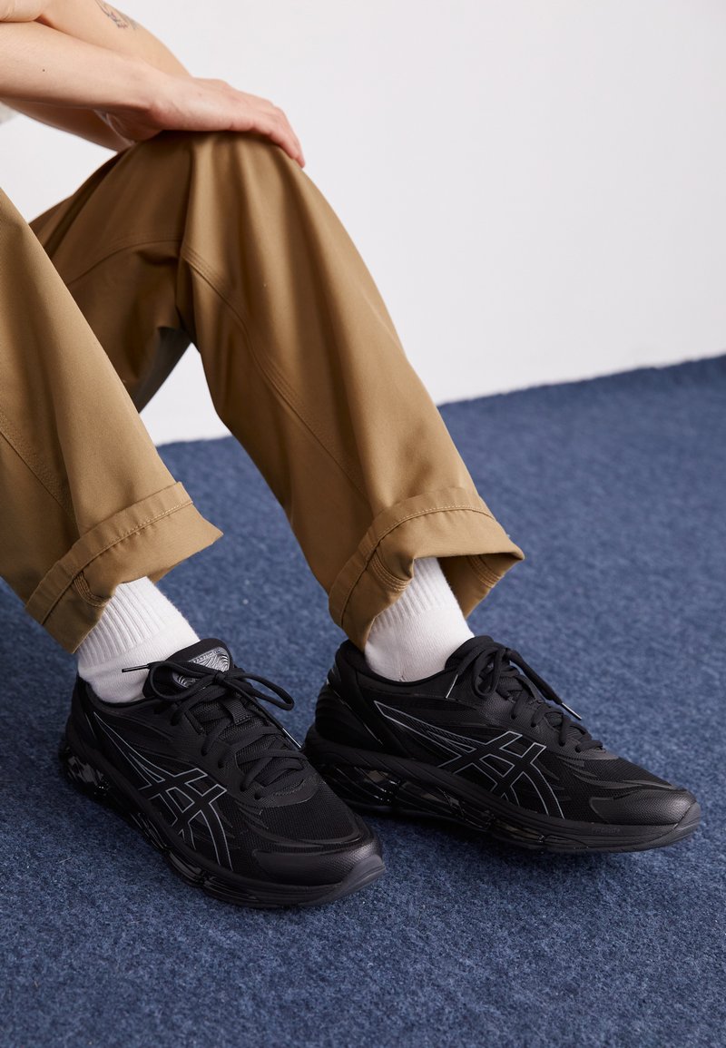 Baskets de course noires avec dessus en mesh, accents réfléchissants et semelle rembourrée, associées à un pantalon beige et des chaussettes blanches montantes sur un tapis bleu.