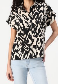 Chemise à manches courtes avec un motif abstrait noir et crème, coupe décontractée et deux poches poitrine. Tissu lisse.