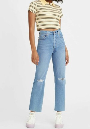 Junge Person trägt ein beiges, gestreiftes, kurzes Poloshirt, hochtaillierte, hellblaue, zerrissene Jeans und weiße Sneakers vor weißem Hintergrund.