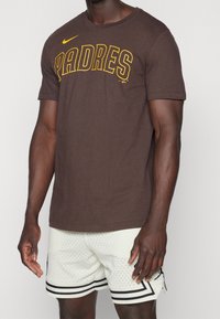 Man som bär en brun "Padres" Nike T-shirt och vita perforerade basketshorts med svarta ränder, stående mot en enfärgad bakgrund.