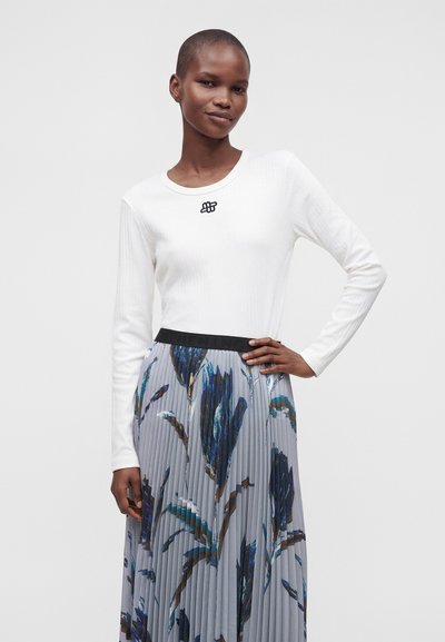 Femme portant une chemise blanche à manches longues avec un petit logo noir et une jupe plissée à motif floral bleu, debout avec une main sur la hanche.