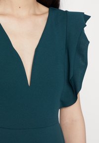 Tyrkysové šaty s hlubokým V-výstřihem, volánovými rukávy a hladkou texturou, s přiléhavým pasem a minimalistickými designovými prvky.