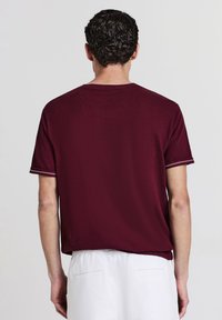 Uomo con capelli ricci scuri che indossa una camicia a maniche corte color bordeaux e pantaloni bianchi, visto da dietro su uno sfondo chiaro.