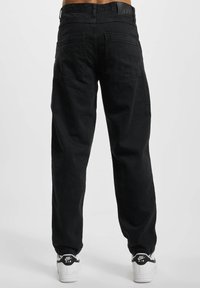 Jeans de mezclilla negra con un corte relajado, dos bolsillos traseros y una etiqueta cosida en la cintura. Combinados con zapatillas blancas que tienen un logo negro.