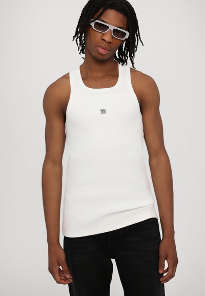 Hvid tanktop med ribbet tekstur, rund halsudskæring og lille grå logo centreret på brystet. Bæres med mørke jeans og briller.