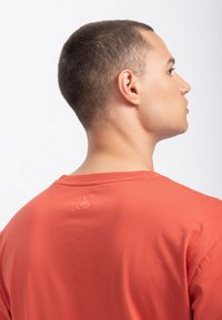 Rode katoenen t-shirt met een ronde halslijn en een kleine fietsbroderie op de achterkant, getoond op een model van opzij.