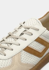 Λευκό δερμάτινο αθλητικό παπούτσι με μπεζ σουέντ λεπτομέρειες, διάτρητο σχέδιο, μπεζ κορδόνια και ανοιχτό καφέ σόλα. Κοντινή όψη των πλευρικών λεπτομερειών.
