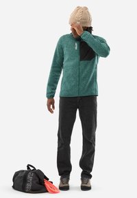 Teal fleece jacket with black chest pocket, paired with dark jeans and beige shoes. Black duffel bag with pink rope nearby.