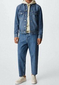 Blauwe spijkerjacket met knoopsluiting en twee borstzakken, gecombineerd met bijpassende losse jeans en beige sneakers op een effen achtergrond.