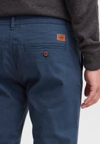 Pantalons en coton bleu marine avec une taille plate, poche arrière et étiquette en cuir marron. Finition avec un détail de bouton marron sur la poche.