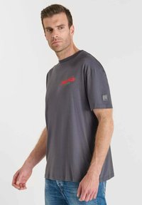 Man in een grijze graphic t-shirt en blauwe spijkerbroek, staande tegen een eenvoudige witte achtergrond, kijkend naar de zijkant.