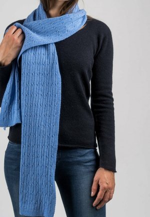 Écharpe tricotée bleu clair avec une texture côtelée, portée sur un pull noir, présentant une forme longue et rectangulaire avec quelques détails subtils.