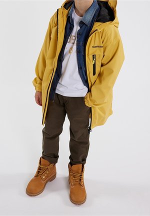 Dziecko w żółtej kurtce przeciwdeszczowej Timberland na koszuli jeansowej i białej koszulce, brązowych spodniach i beżowych sznurowanych butach na jednolitym tle.