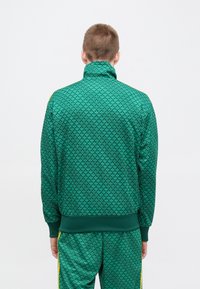 Πράσινο μπουφάν φόρμας με ψηλό γιακά, υφασμένο σχέδιο με στυλιζαρισμένα φύλλα και ριμπ τελειώματα στα μανίκια και το τελείωμα. Περιλαμβάνει κίτρινες πλευρικές λεπτομέρειες.