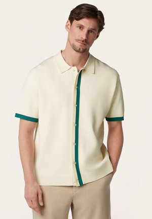 Man draagt een korte mouw crèmeoverhemd met groene bies op de knoopsluiting en mouwboorden, gecombineerd met beige broek, poserend tegen een effen achtergrond.