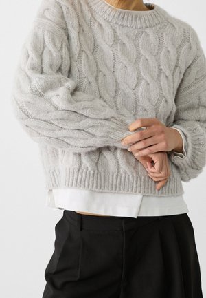 Persoon draagt een lichtgrijze kabelgebreide trui over een wit shirt, met een zwarte broek, handen zachtjes één pols vasthoudend.