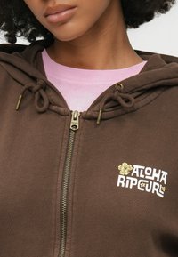 Καφέ φούτερ με φερμουάρ και ρυθμιζόμενα κορδόνια, που έχει χρυσό φερμουάρ, κεντημένο λευκό και κίτρινο λογότυπο "ALOHARIP CURL", φορεμένο πάνω από ροζ μπλουζάκι.