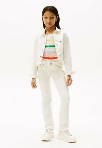 Weiße Denim-Jacke über einem mehrfarbigen, gemusterten Pullover, kombiniert mit weißen Skinny-Jeans und weißen Sneakers. Einfaches Design und neutrale Farben.