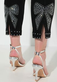 Černé denní kalhoty s velkými designy mašlí z rhinestone na nohavicích, zdobené lemy a stříbrné sandály na vysokém podpatku s kotníkovými pásky.