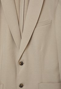 Blazer beige con una trama strutturata, due bottoni scuri, rever a punta e una tasca sul petto.