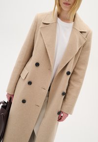 Bežs vilnas sakausējuma mētelis ar divkāršo pogu dizainu, lieliem atloka galiem un melnām pogām. Iezīmē sānu kabatas un gludu tekstūru.