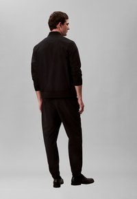 Mees seisab seljaga vaataja poole, kandes musta jope, musti pükse ja musti kingi ning neid ümbritseb hall, monokroomne taust.