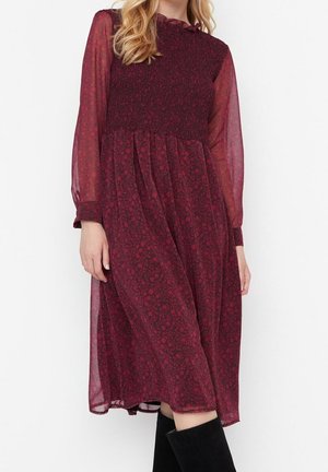 Femme portant une robe midi florale bordeaux à manches longues, avec un corsage smocké et des manches transparentes, associée à des bottes noires montantes jusqu'aux genoux.
