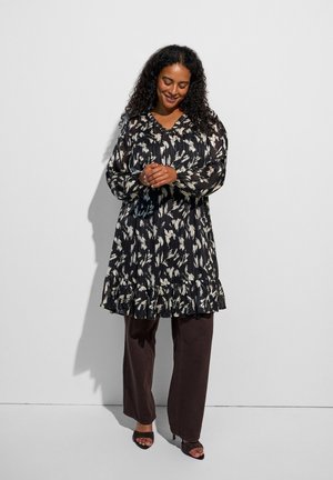 Donna con capelli ricci che indossa un vestito a maniche lunghe con motivo bianco e nero sopra pantaloni scuri e tacchi neri aperti sulla punta, in piedi contro un muro bianco.